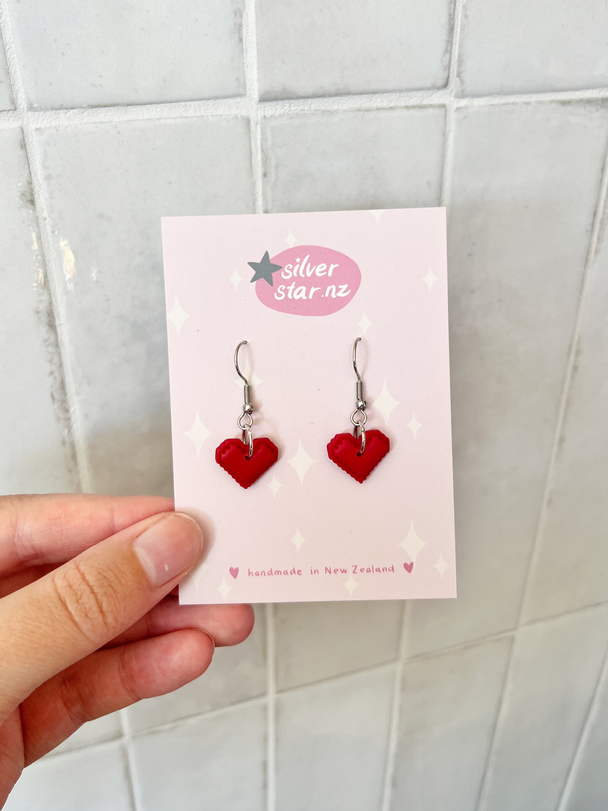 A hand holds a card labeled silverstar.nz with Pixel Heart Polymer Clay Earrings—red heart-shaped handmade pieces on stainless steel hooks, crafted in New Zealand, shown against a tiled background.