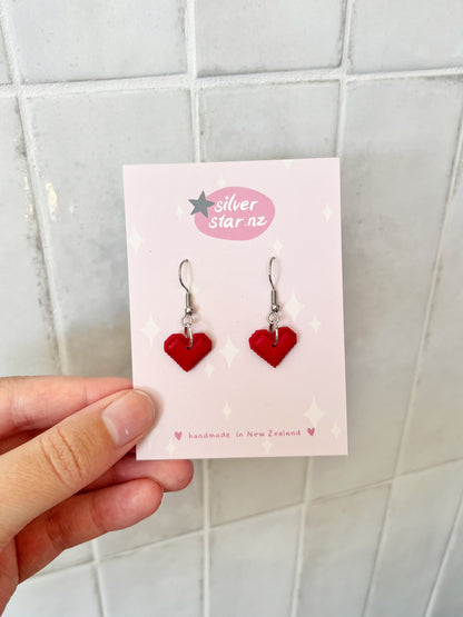 A hand holds a card labeled silverstar.nz with Pixel Heart Polymer Clay Earrings—red heart-shaped handmade pieces on stainless steel hooks, crafted in New Zealand, shown against a tiled background.