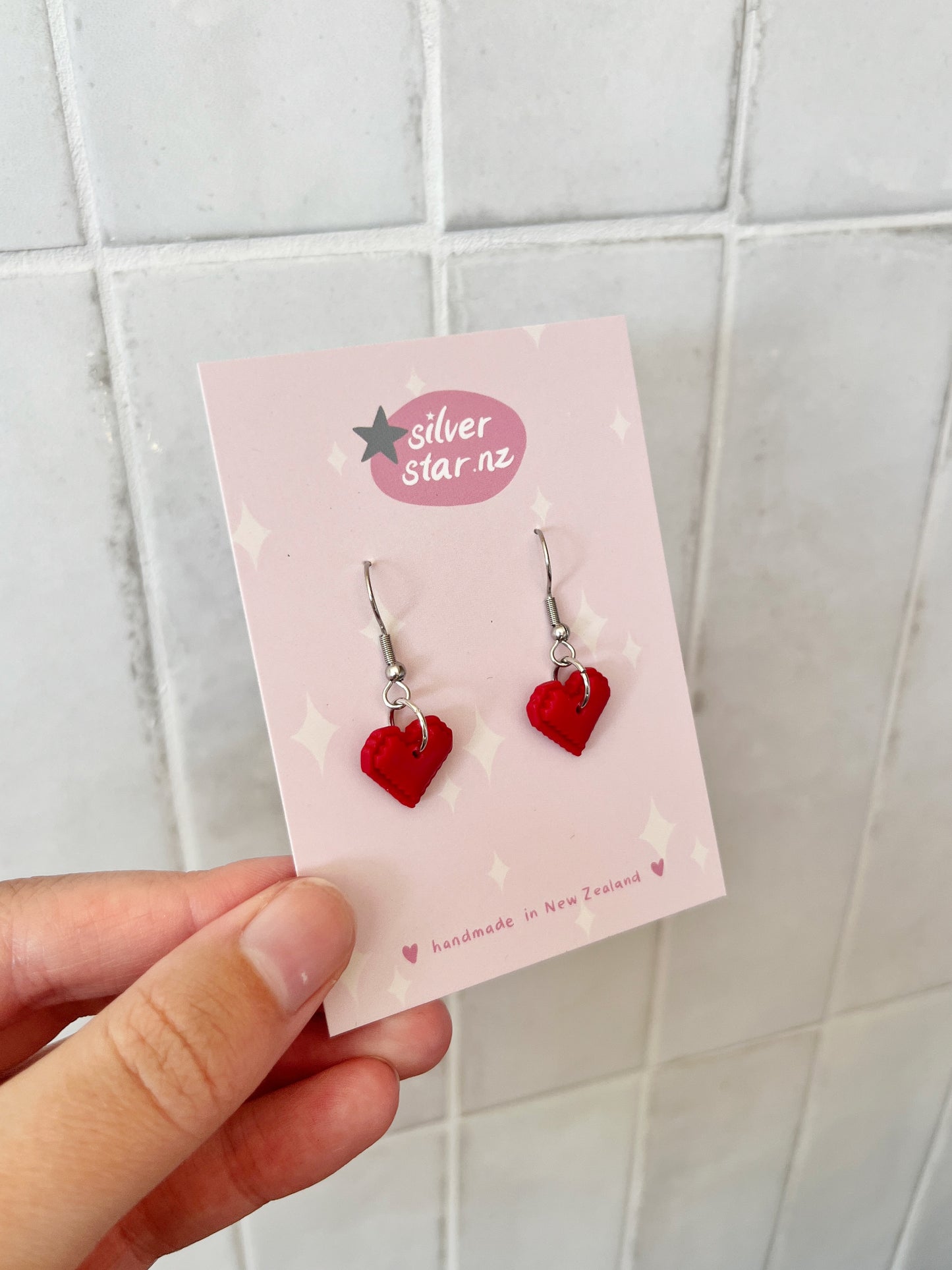A hand holds a pink card with white stars showing Pixel Heart Polymer Clay Earrings and the silverstar.nz logo. The card reads Handmade in New Zealand and displays red heart-shaped earrings with stainless steel hooks.