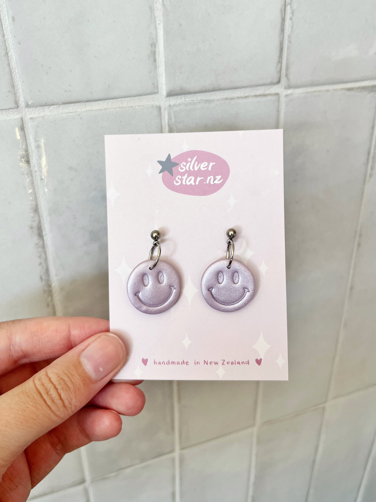 A hand holds a card displaying Smiley Polymer Clay Earrings by silverstar.nz, with “silverstar.nz” at the top and “handmade in New Zealand” at the bottom. The background features light tiles.