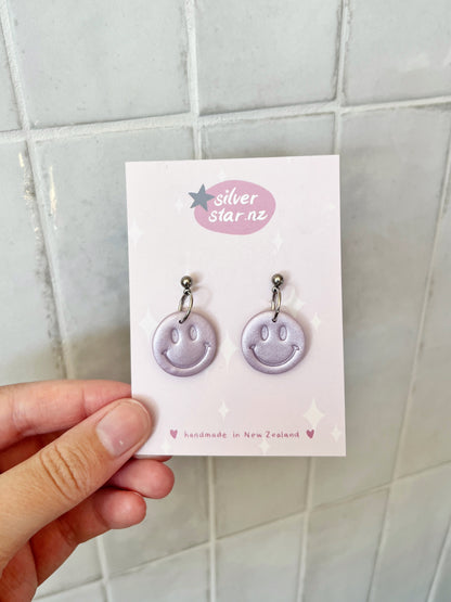 A hand holds a card displaying Smiley Polymer Clay Earrings by silverstar.nz, with “silverstar.nz” at the top and “handmade in New Zealand” at the bottom. The background features light tiles.