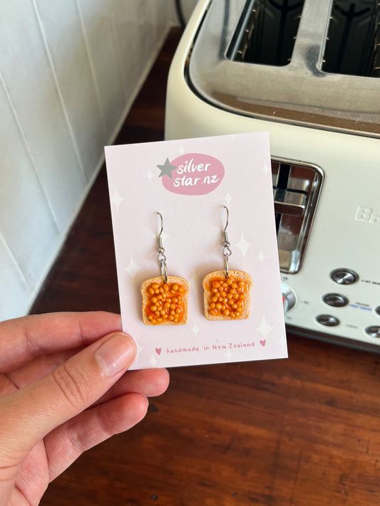 A hand holds a card with silverstar.nz Beans on Toast Polymer Clay Earrings, designed to look like toast topped with orange beans. A white toaster rests in the background on a wooden countertop.
