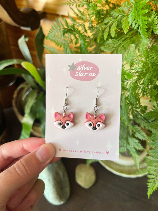 A hand displays pink fox-shaped earrings made from handmade polymer clay on a Silver Star NZ card. Surrounded by green houseplants, stones, and wood, they are labeled as handmade in New Zealand with a unique funky design. Product: Fox Polymer Clay Earrings by silverstar.nz.