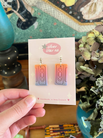 A hand displays Casette Polymer Clay Earrings by silverstar.nz, shaped like colorful cassette tapes in pink and purple gradients. The card says “silverstar.nz” and “handmade in New Zealand,” with flowers and decor in the background.