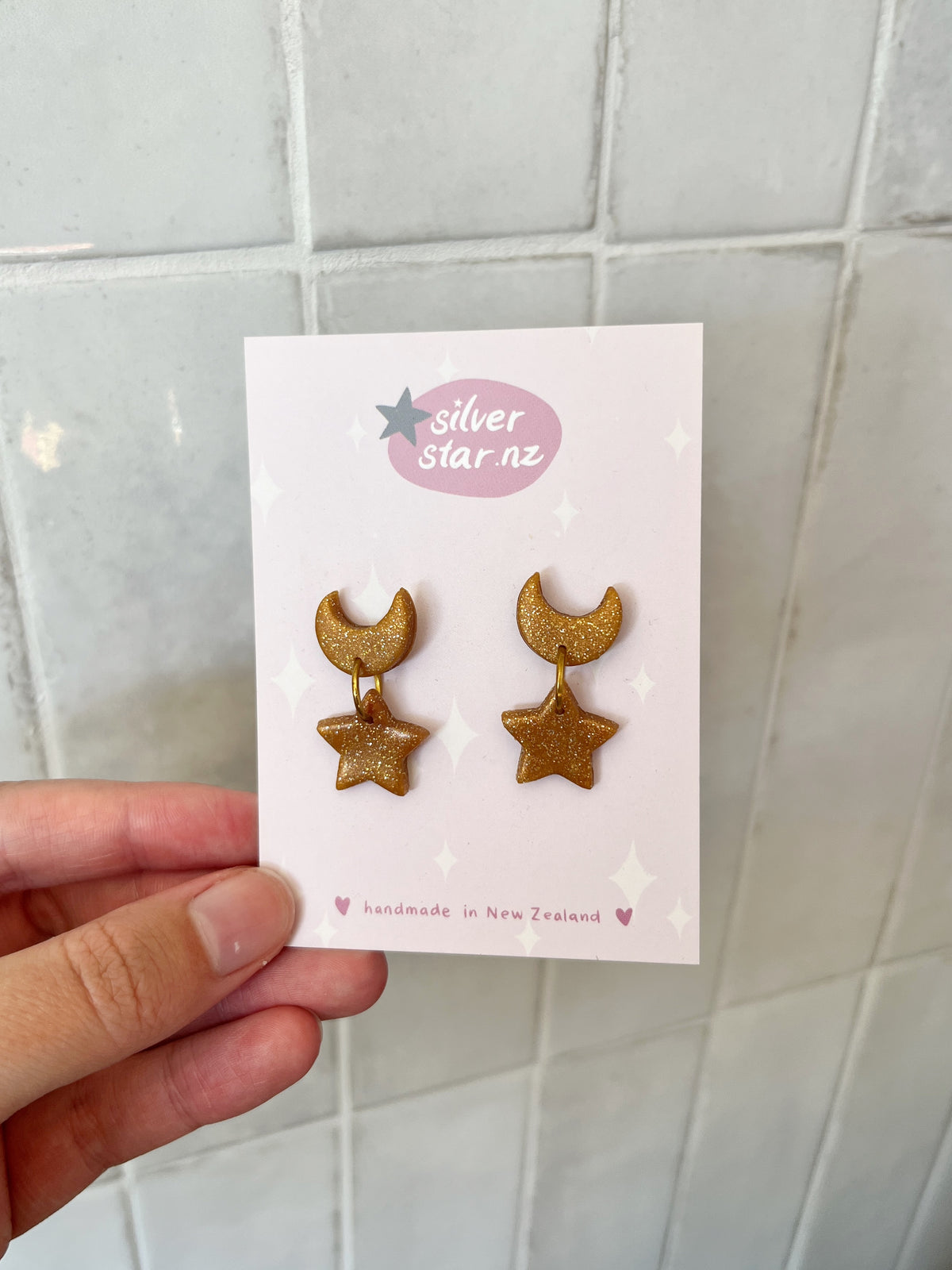 A hand holds a card with Star + Moon Polymer Clay Earrings featuring unique gold glitter crescent moon and star designs. The card reads "silverstar.nz" and "handmade in New Zealand," surrounded by decorative stars.