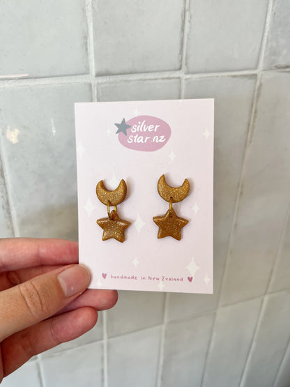 A hand holds a card with Star + Moon Polymer Clay Earrings featuring unique gold glitter crescent moon and star designs. The card reads "silverstar.nz" and "handmade in New Zealand," surrounded by decorative stars.
