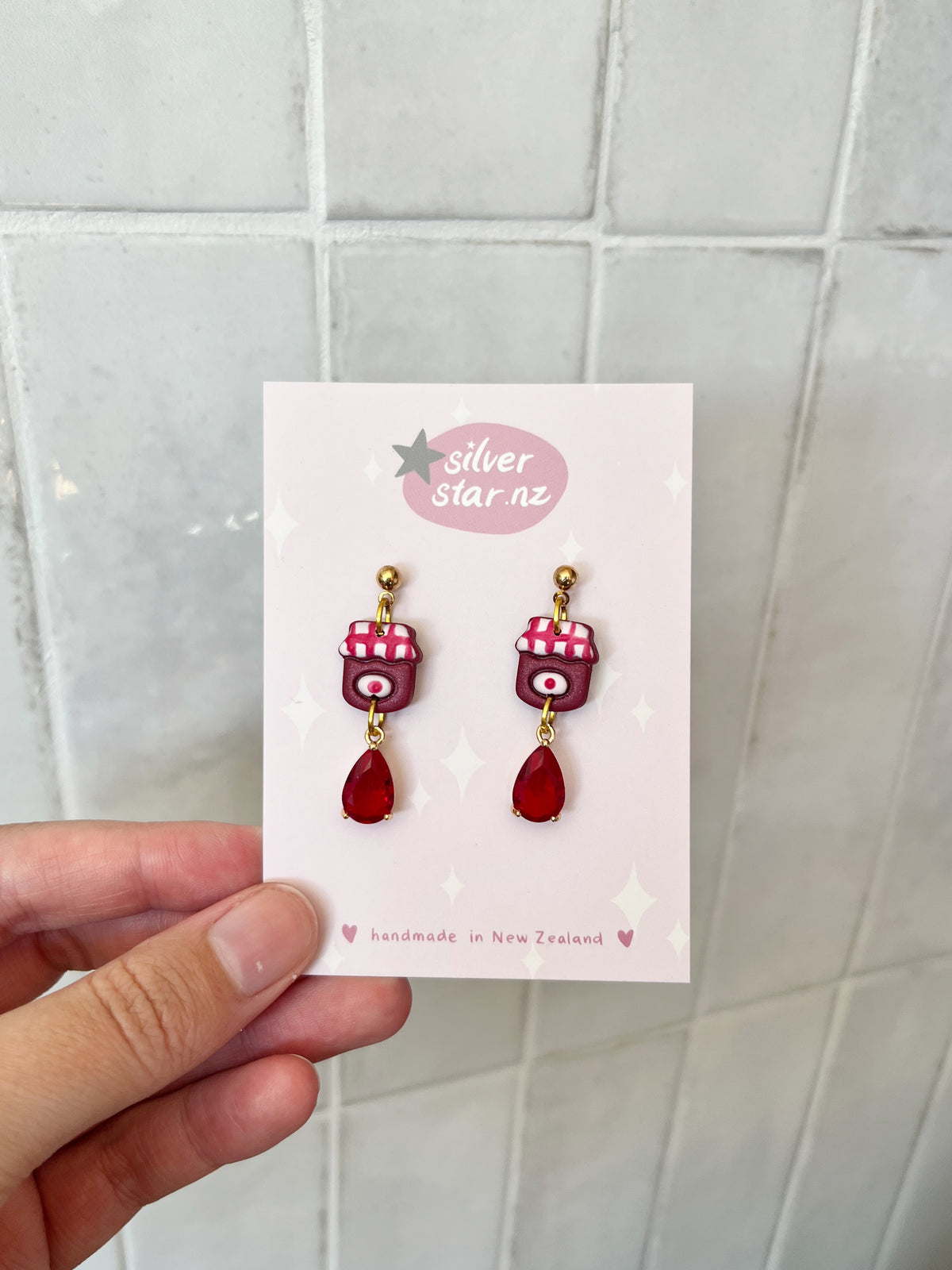 A hand holds a card displaying "Berry Jam Jar Polymer Clay Earrings" by silverstar.nz, featuring handmade polymer clay charms and stainless steel hooks, labeled handmade in New Zealand against a tiled background.