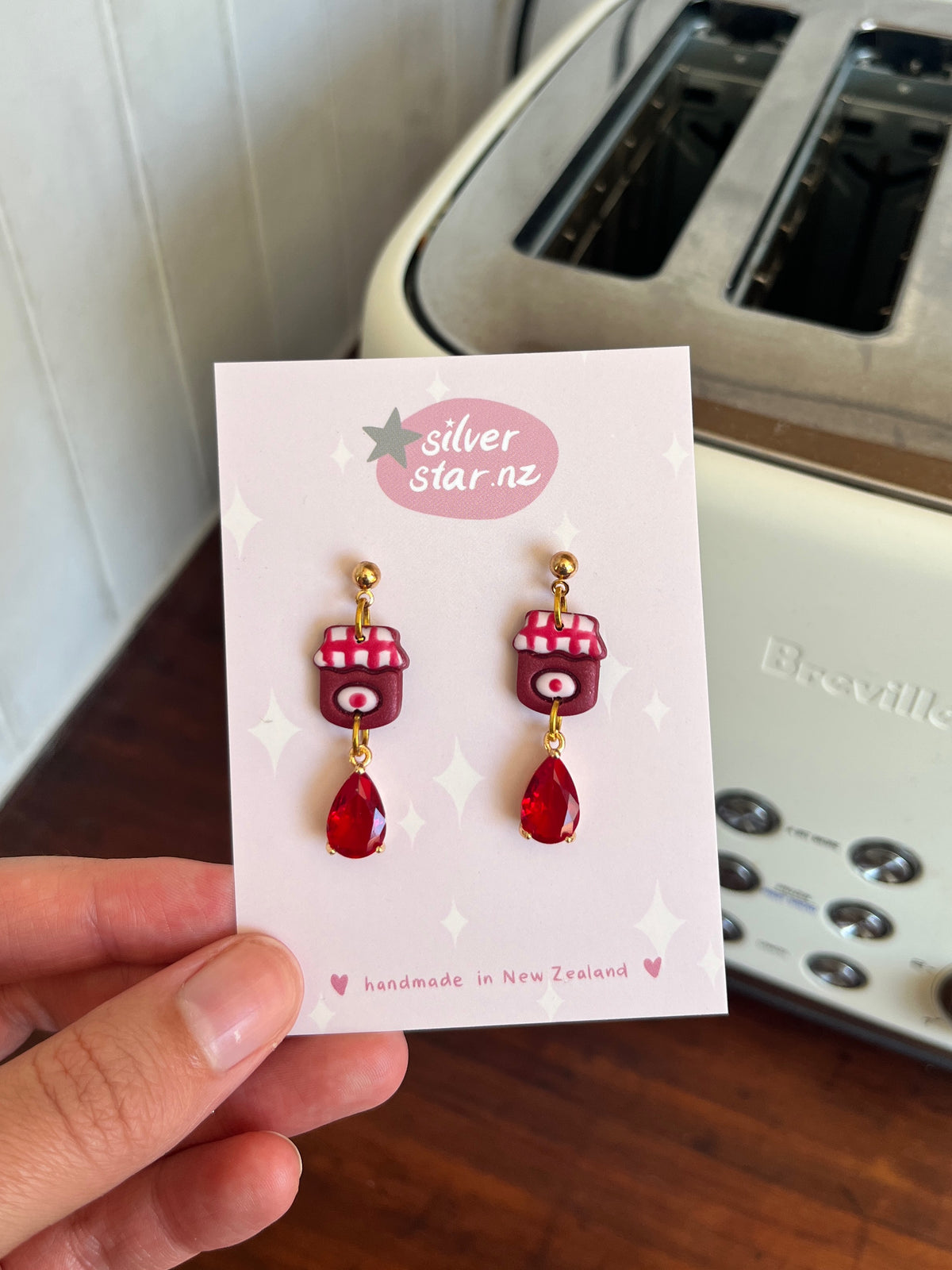 A hand holds a card reading silverstar.nz with Berry Jam Jar Polymer Clay Earrings, handmade in New Zealand. In the background is a white Breville toaster.