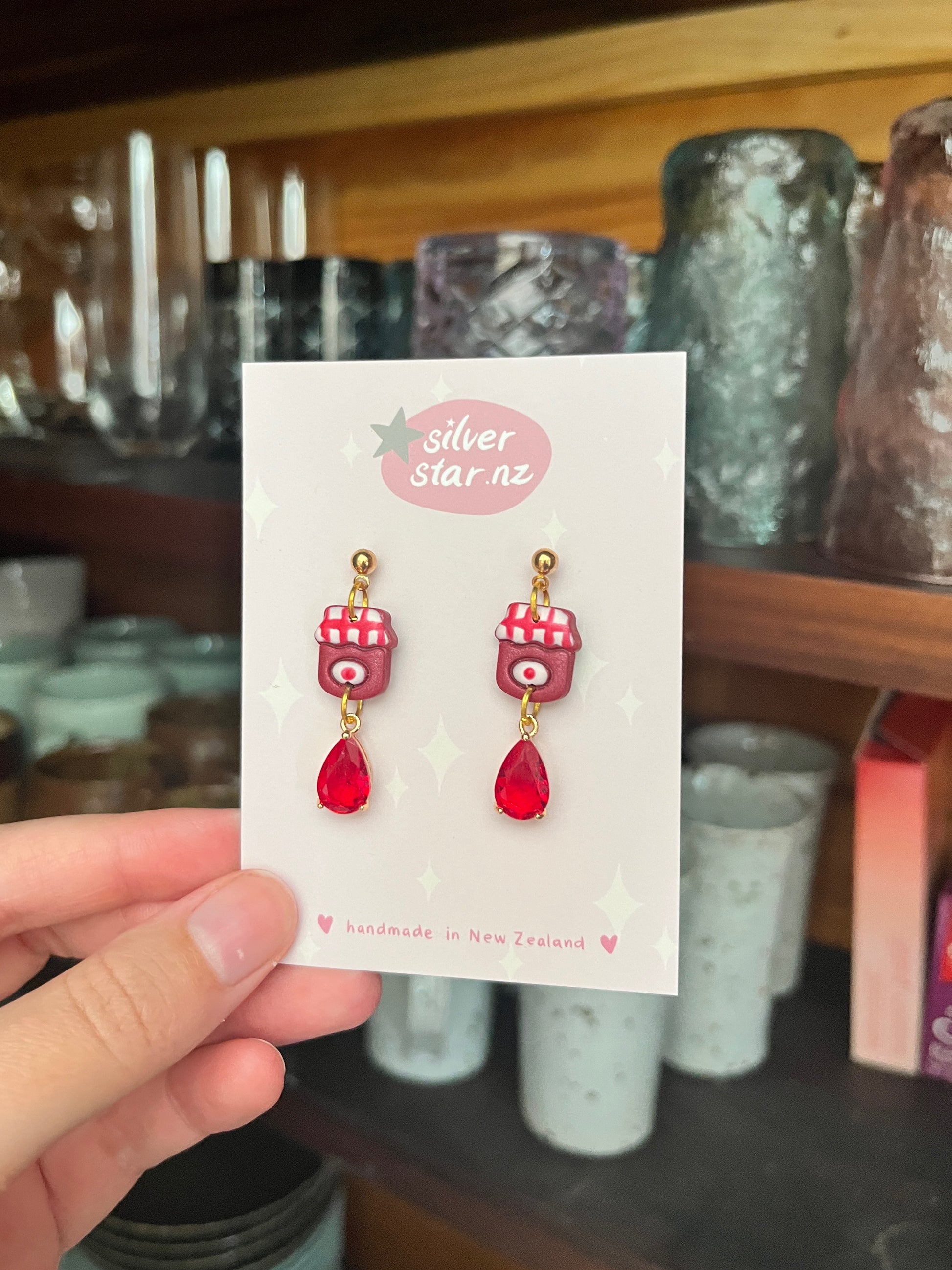 A hand holds a card displaying Berry Jam Jar Polymer Clay Earrings by silverstar.nz, handmade in New Zealand, with shelves of glassware visible in the background.