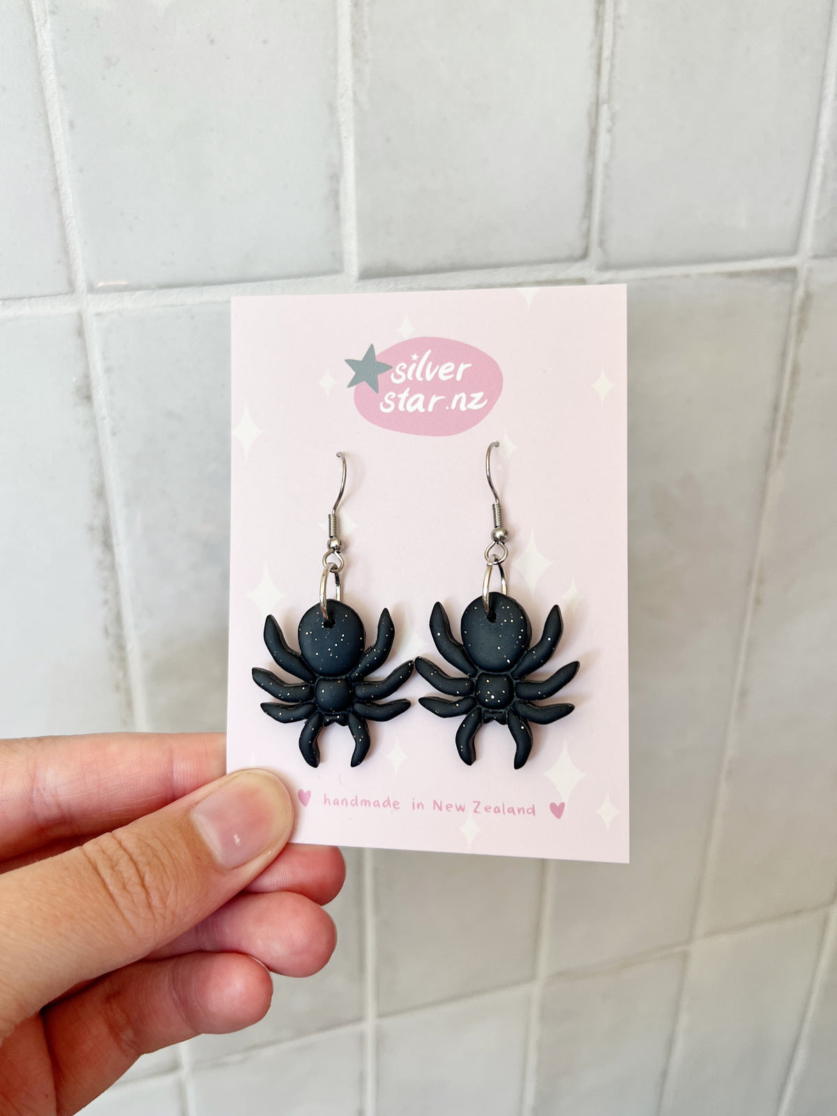 A hand holds a card with Spider Polymer Clay Earrings—black, spider-shaped, with stainless steel hooks. The card reads silverstar.nz and handmade in New Zealand, shown against a tiled background.