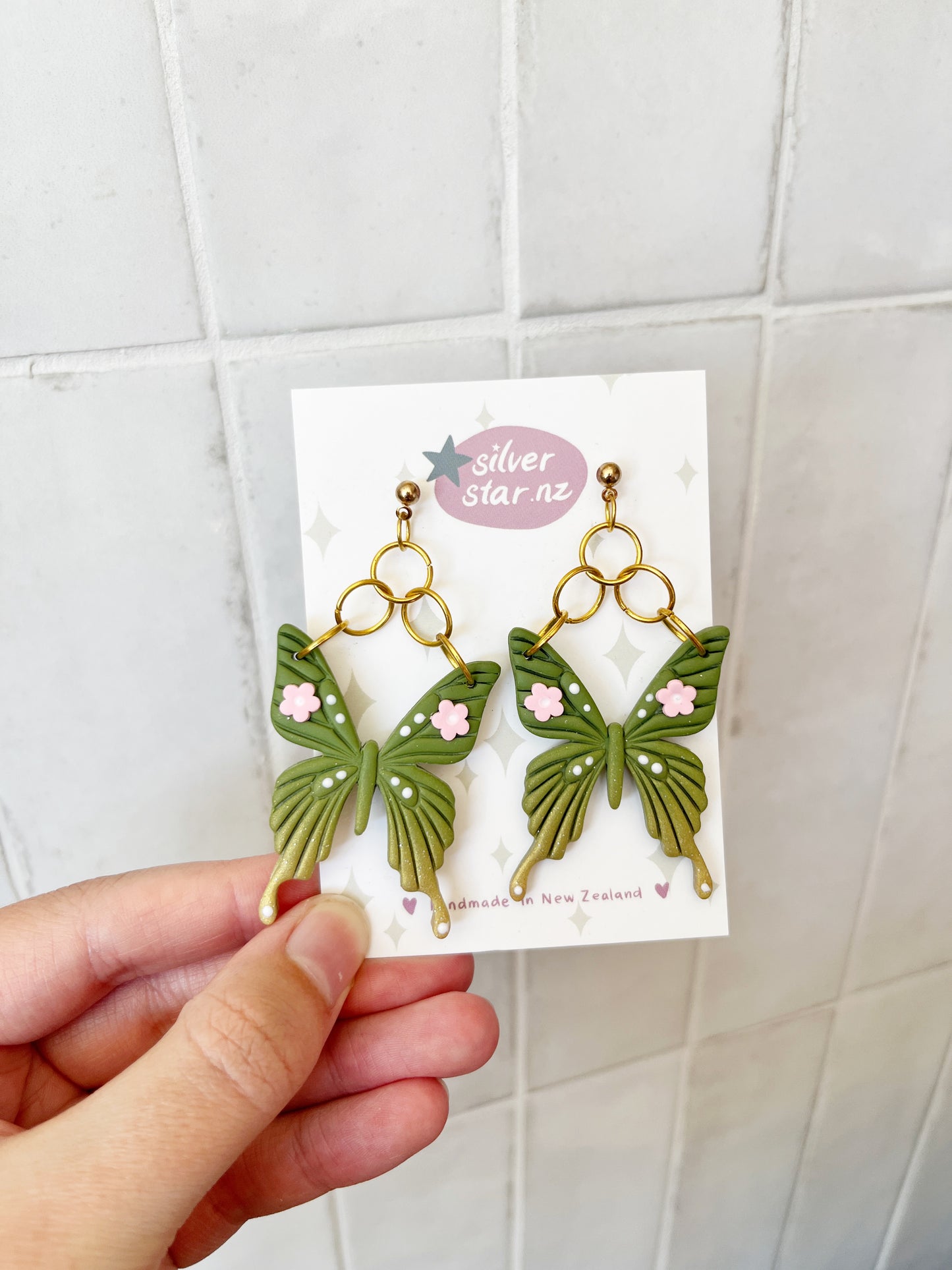 A hand holds a card displaying Green Fae Butterfly Polymer Clay Earrings by silverstar.nz—green butterfly shapes with pink flowers and gold loops. The card says "Handmade in New Zealand" on a tiled background.