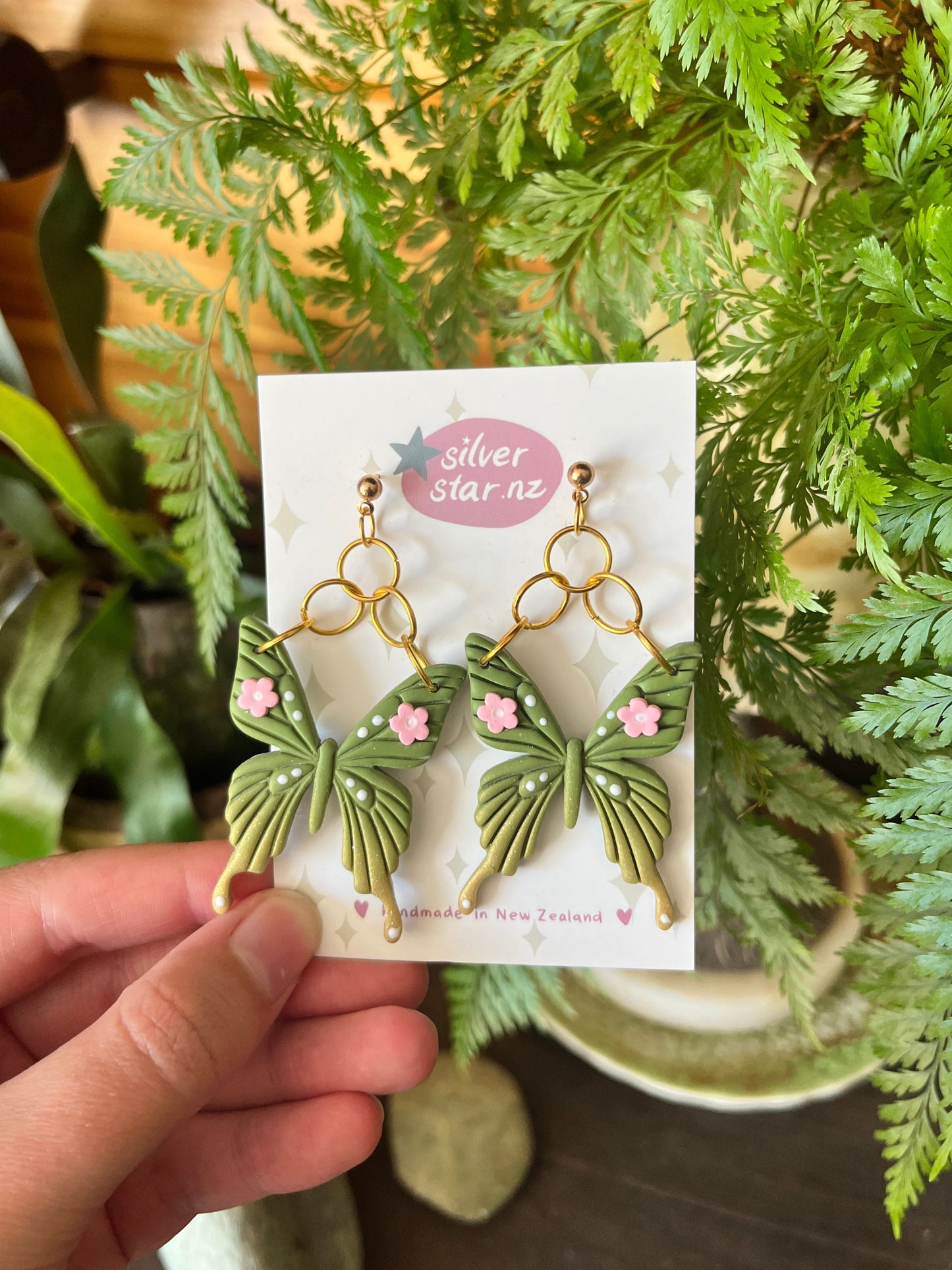 A hand holds a card with Green Fae Butterfly Polymer Clay Earrings by silverstar.nz—green butterfly shapes with tiny pink flowers and stainless steel hooks, displayed against lush green plants.