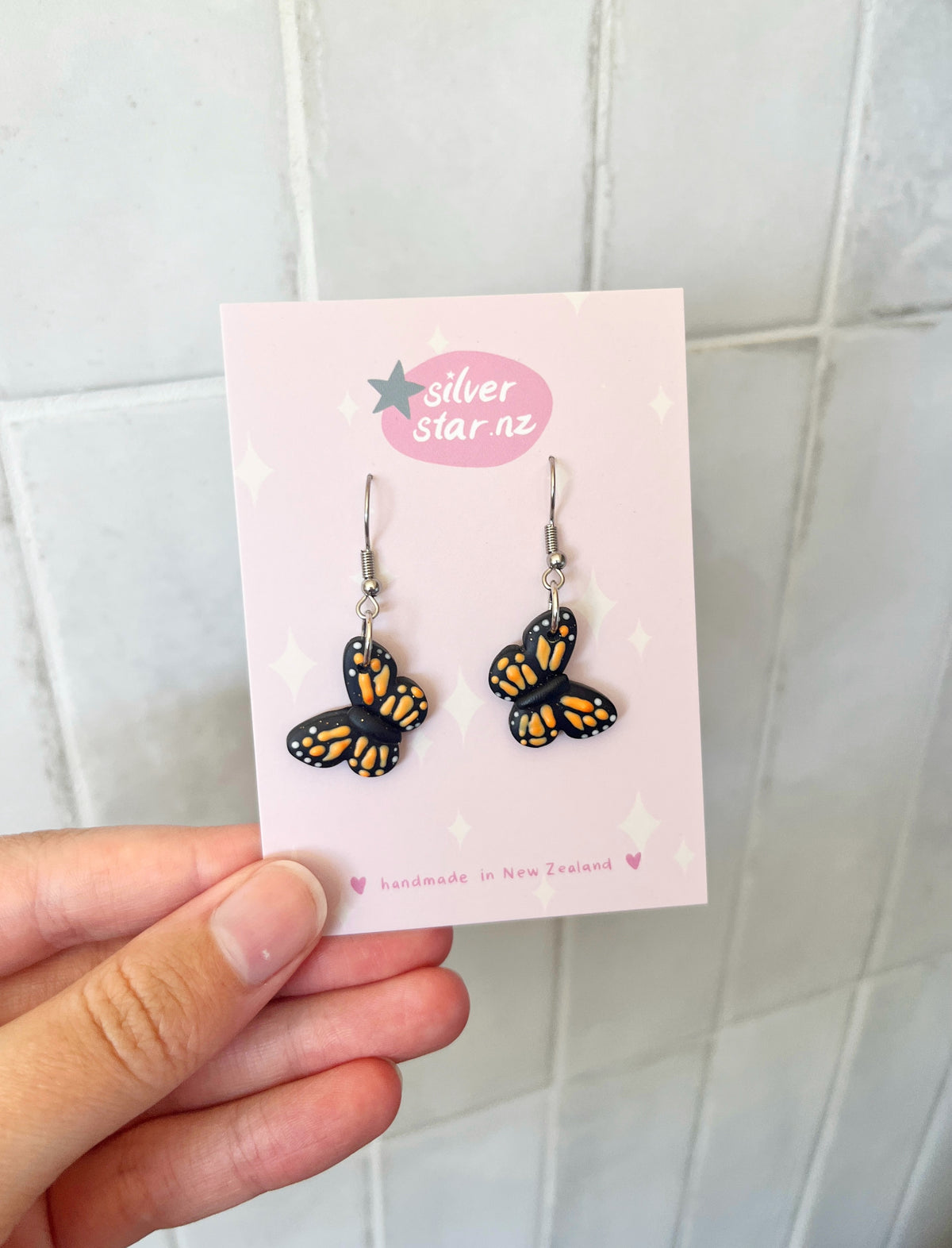 A hand holds a card exhibiting Monarch Butterfly Polymer Clay Earrings from silverstar.nz. These black and orange butterfly-shaped earrings with stainless steel hooks resemble monarchs and rest on a pink background with tiny stars, stating silver star nz and handmade in New Zealand.