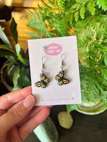 A hand displays Monarch Butterfly Polymer Clay Earrings with black and orange details on a silverstar.nz, handmade in New Zealand card. These unique earrings feature stainless steel hooks, set against a backdrop of lush foliage and smooth stones.