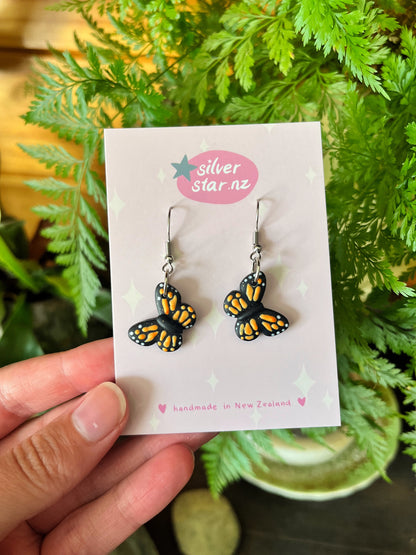 A hand displays the Monarch Butterfly Polymer Clay Earrings, featuring a black design with orange and white accents. The durable earrings have stainless steel hooks, and the card reads silverstar.nz and handmade in New Zealand against a leafy background.