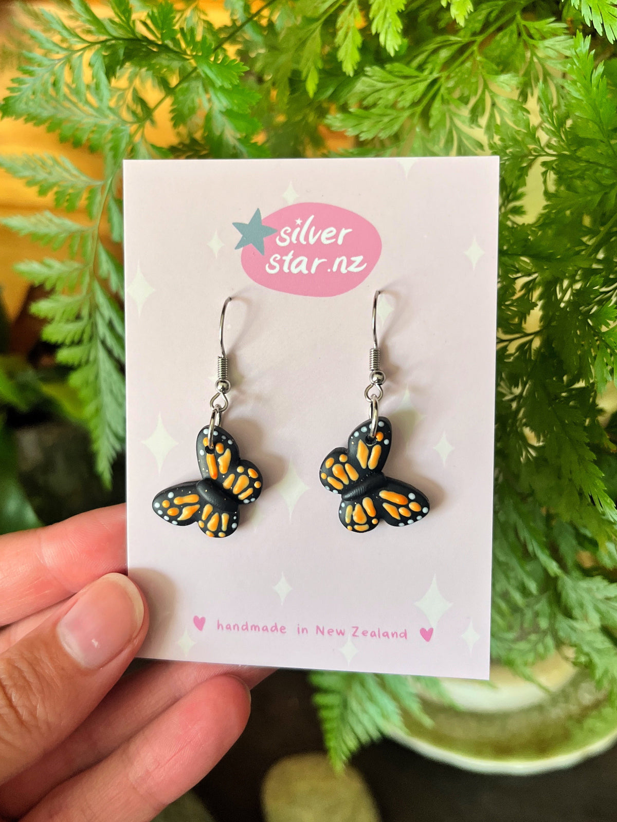 Monarch Butterfly Polymer Clay Earrings by silverstar.nz feature a unique butterfly design with orange and black wings, stainless steel hooks, and are displayed on a card against green foliage. The text reads handmade in New Zealand.