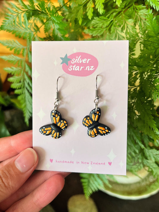 Monarch Butterfly Polymer Clay Earrings by silverstar.nz feature a unique butterfly design with orange and black wings, stainless steel hooks, and are displayed on a card against green foliage. The text reads handmade in New Zealand.