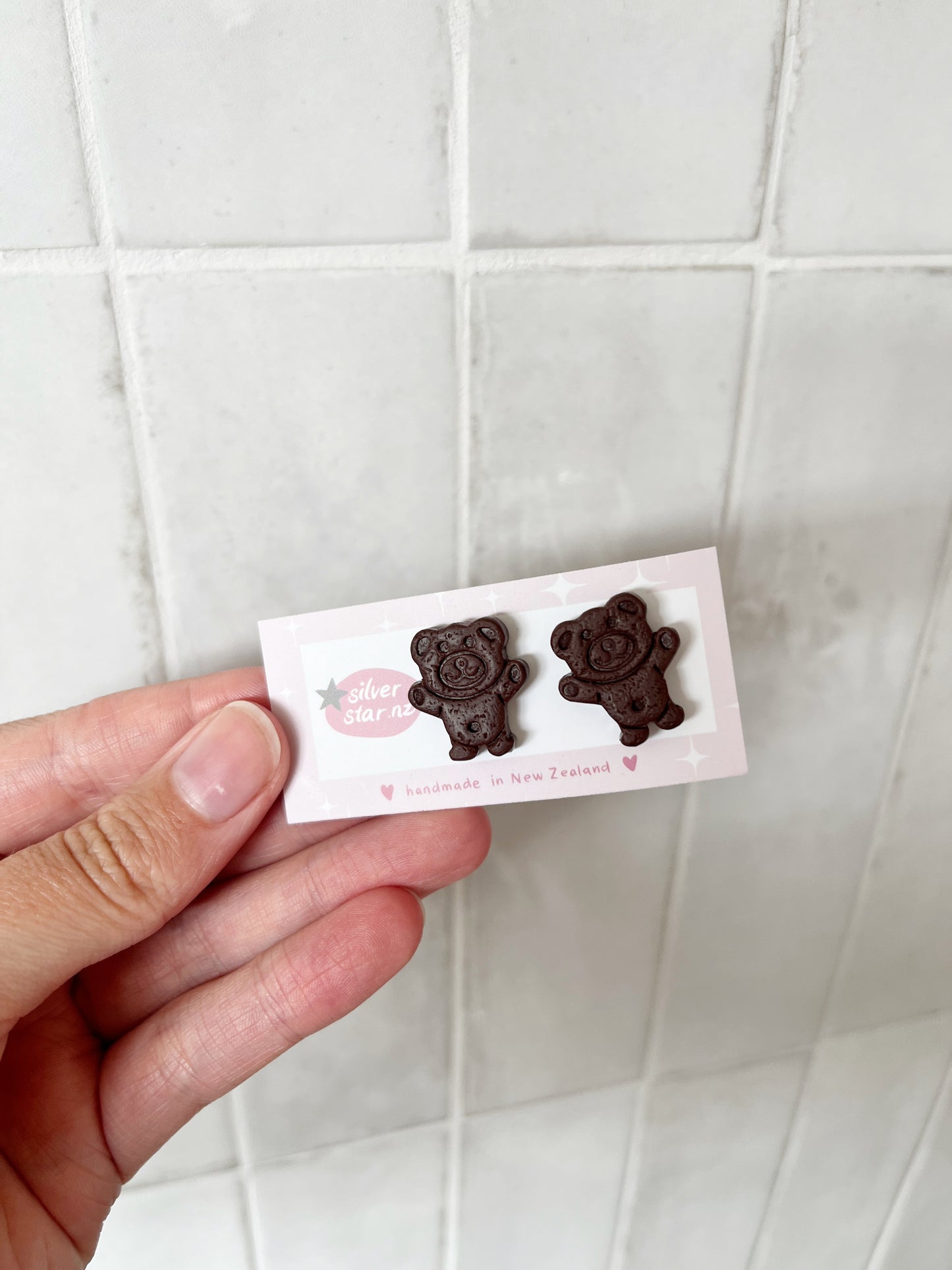 A hand holds a card with two Tiny Teddy Polymer Clay Stud Earrings by silverstar.nz. The card says handmade in New Zealand and is set against a white tiled background.