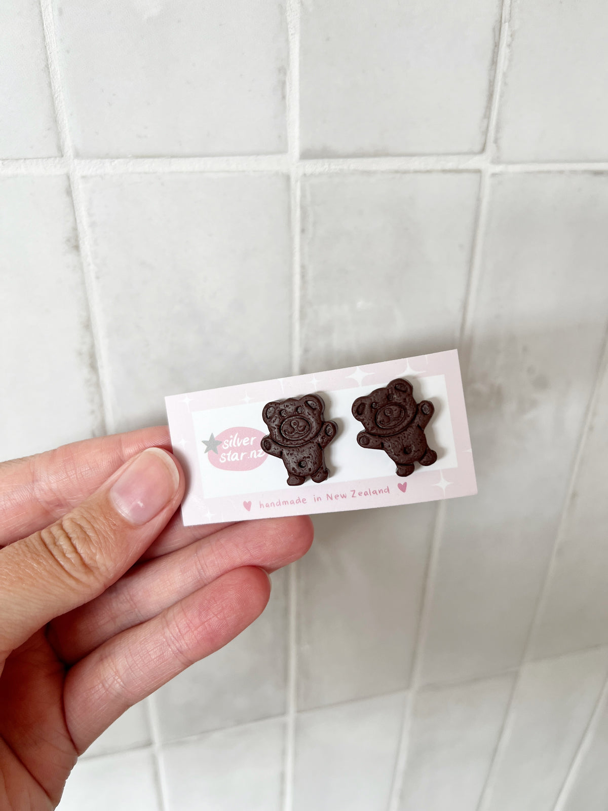 A hand holds a card with two Tiny Teddy Polymer Clay Stud Earrings by silverstar.nz. The card says handmade in New Zealand and is set against a white tiled background.