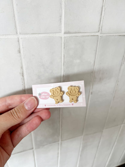 A hand holds a card displaying two gold Tiny Teddy Polymer Clay Stud Earrings by silverstar.nz against a white tiled background. The card also notes these earrings are handmade in New Zealand.