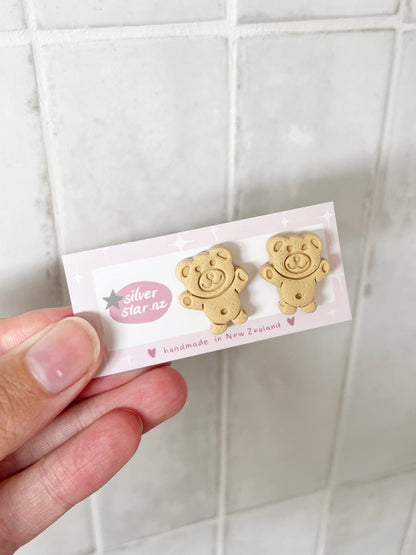 A hand holds a card with Tiny Teddy Polymer Clay Stud Earrings by silverstar.nz, handmade in New Zealand. The earrings resemble cookie-shaped bears. The backdrop is a white tiled wall.