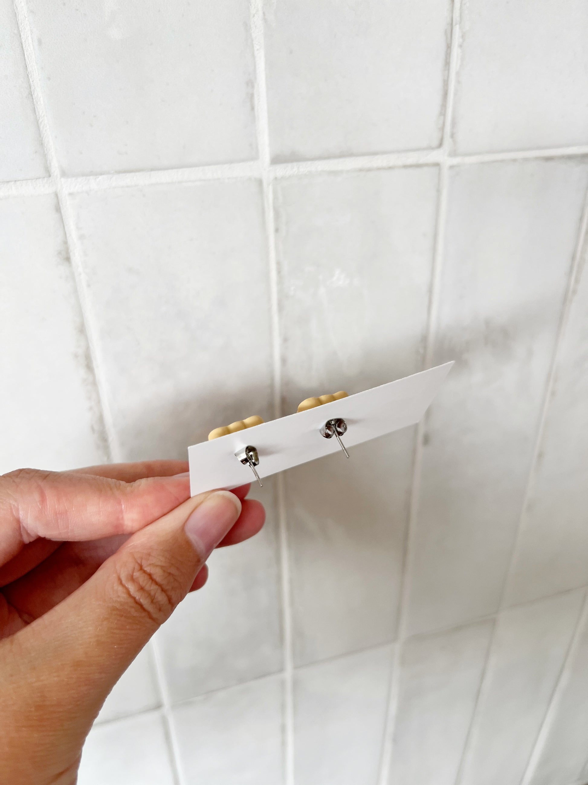 A hand holds a white card with silverstar.nz Tiny Teddy Polymer Clay Stud Earrings (silver posts) in front of a white tiled wall. The front design of these handmade earrings isnt visible.