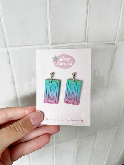 A hand holds a card from silverstar.nz displaying Casette Polymer Clay Earrings in teal, purple, and pink gradients. The card reads “silverstar.nz” and “handmade in New Zealand” with star decorations.