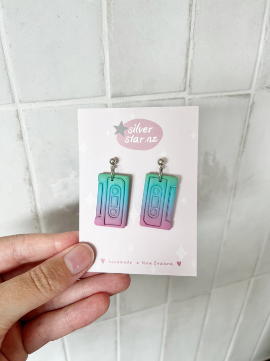 A hand holds a card from silverstar.nz displaying Casette Polymer Clay Earrings in teal, purple, and pink gradients. The card reads “silverstar.nz” and “handmade in New Zealand” with star decorations.