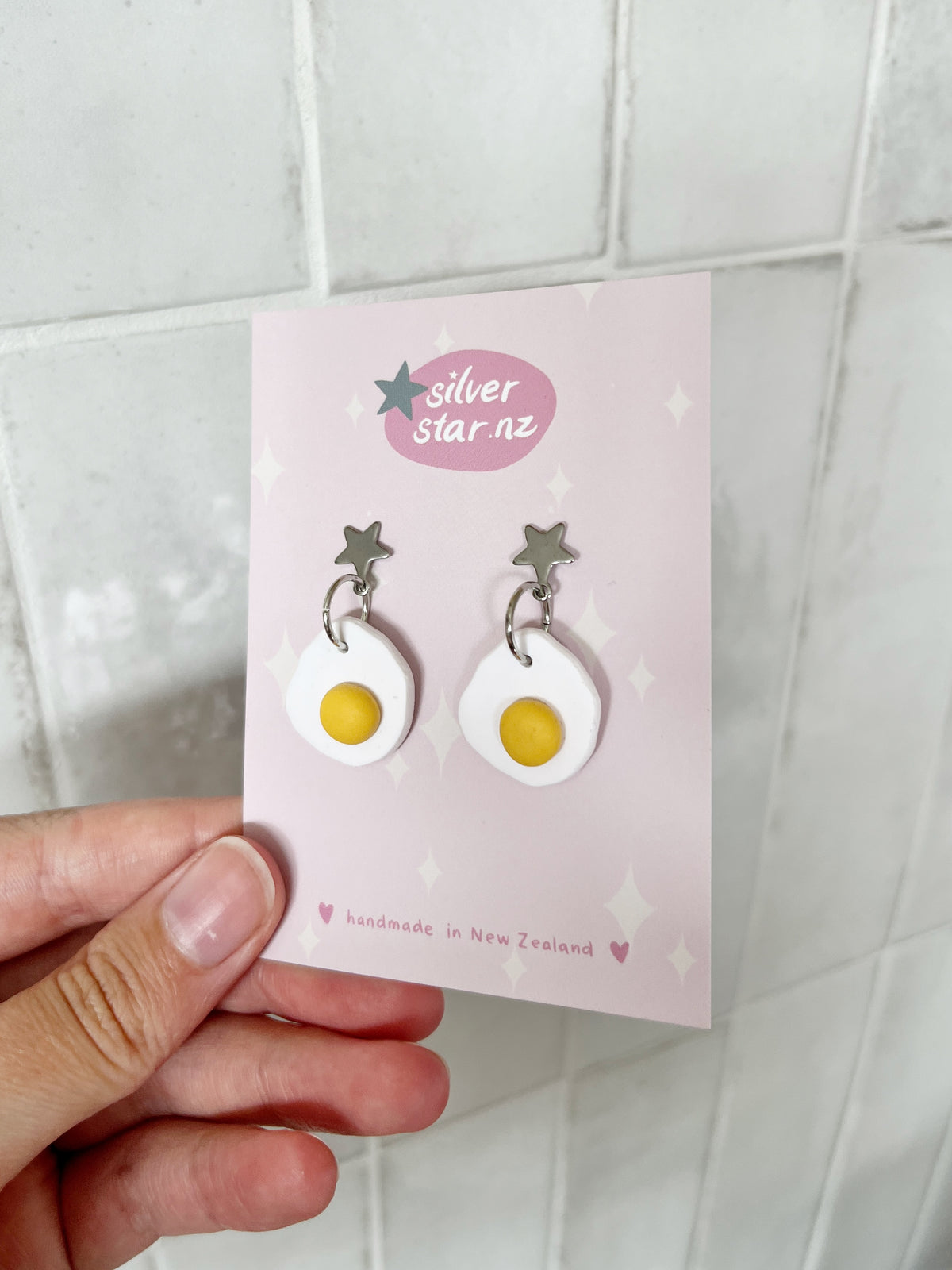 A hand displays a card from silverstar.nz featuring Egg Polymer Clay Earrings—handmade in New Zealand—with star and heart decorations.