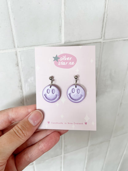 A hand holds a card that reads silverstar.nz and handmade in New Zealand, displaying Smiley Polymer Clay Earrings with lavender smiley faces, star decorations, and stainless steel hooks on a white tiled background.