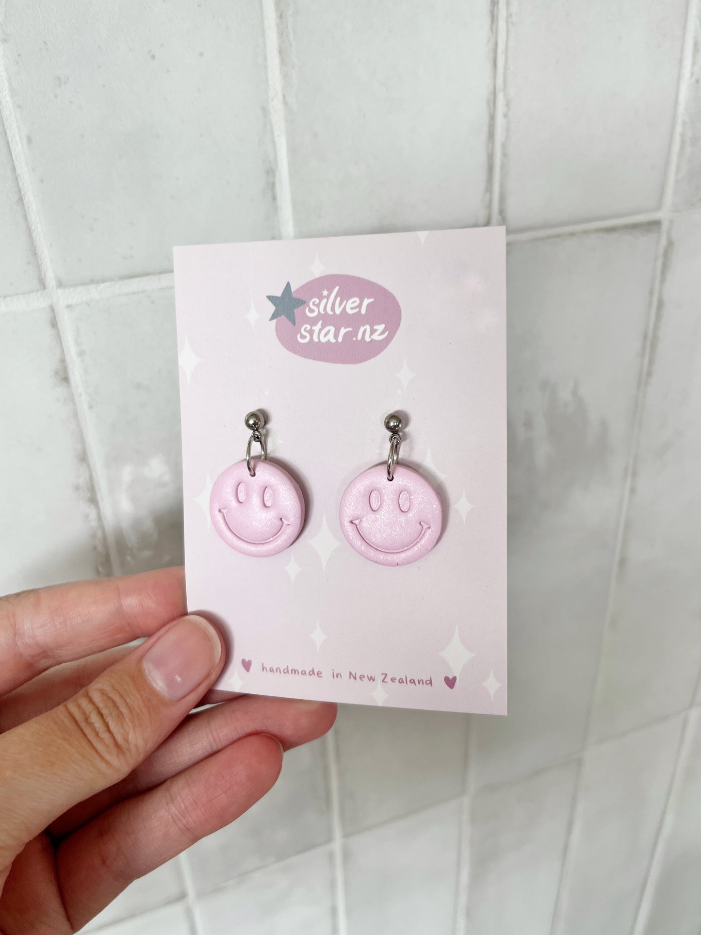 A hand holds a card reading silverstar.nz with Smiley Polymer Clay Earrings—lightweight, handmade in New Zealand, star and sparkle designs, stainless steel hooks—against a light-tiled background.