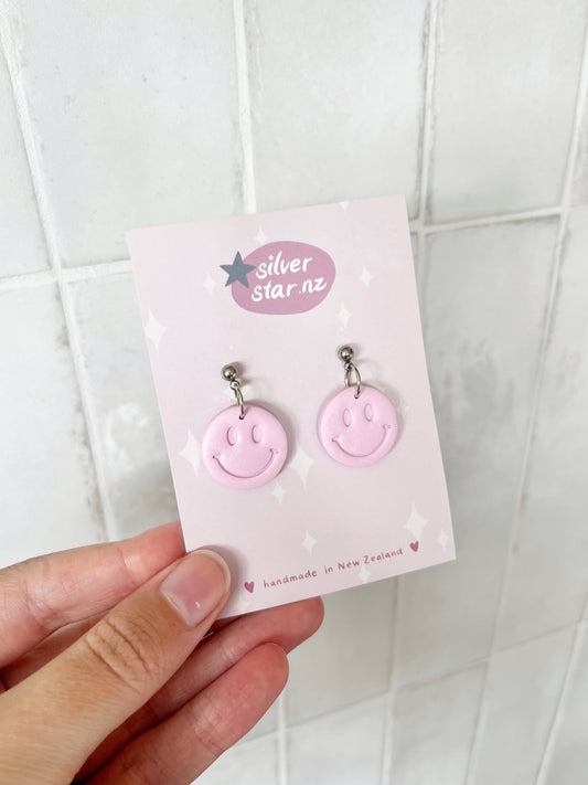 A hand holds a card featuring Smiley Polymer Clay Earrings from silverstar.nz, with handmade in New Zealand at the bottom. Light-colored tiles form the background.