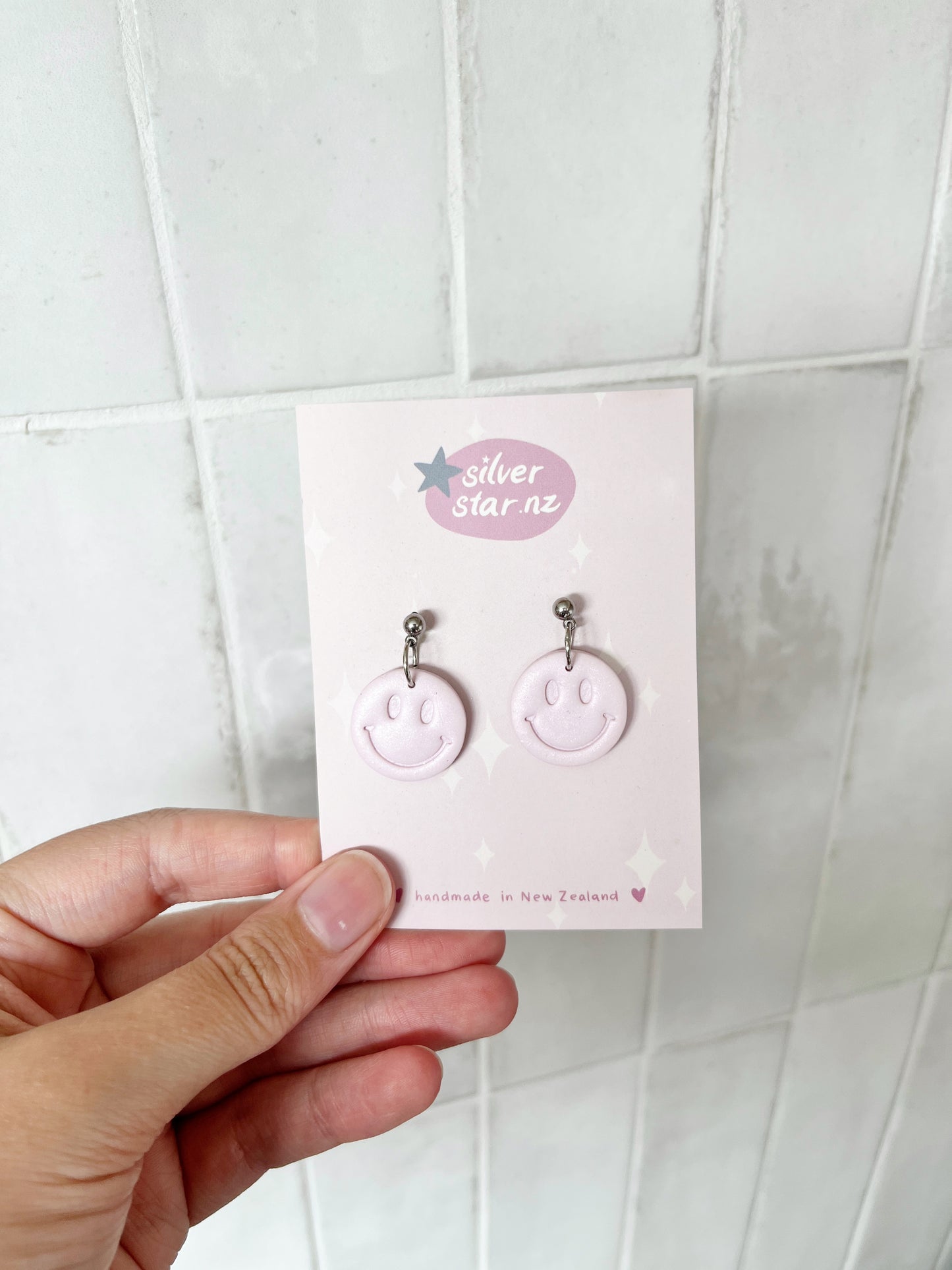 A hand holds a card showing Smiley Polymer Clay Earrings with stainless steel hooks. The card reads silverstar.nz handmade in New Zealand, set against a white tiled background.