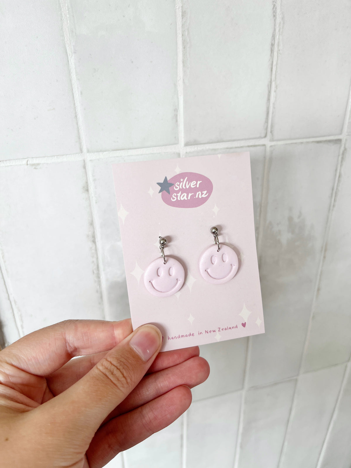 A hand holds a card with Smiley Polymer Clay Earrings—light pink, handmade in New Zealand by silverstar.nz with stainless steel hooks. The card shows the brand’s star logo. White tiles are in the background.