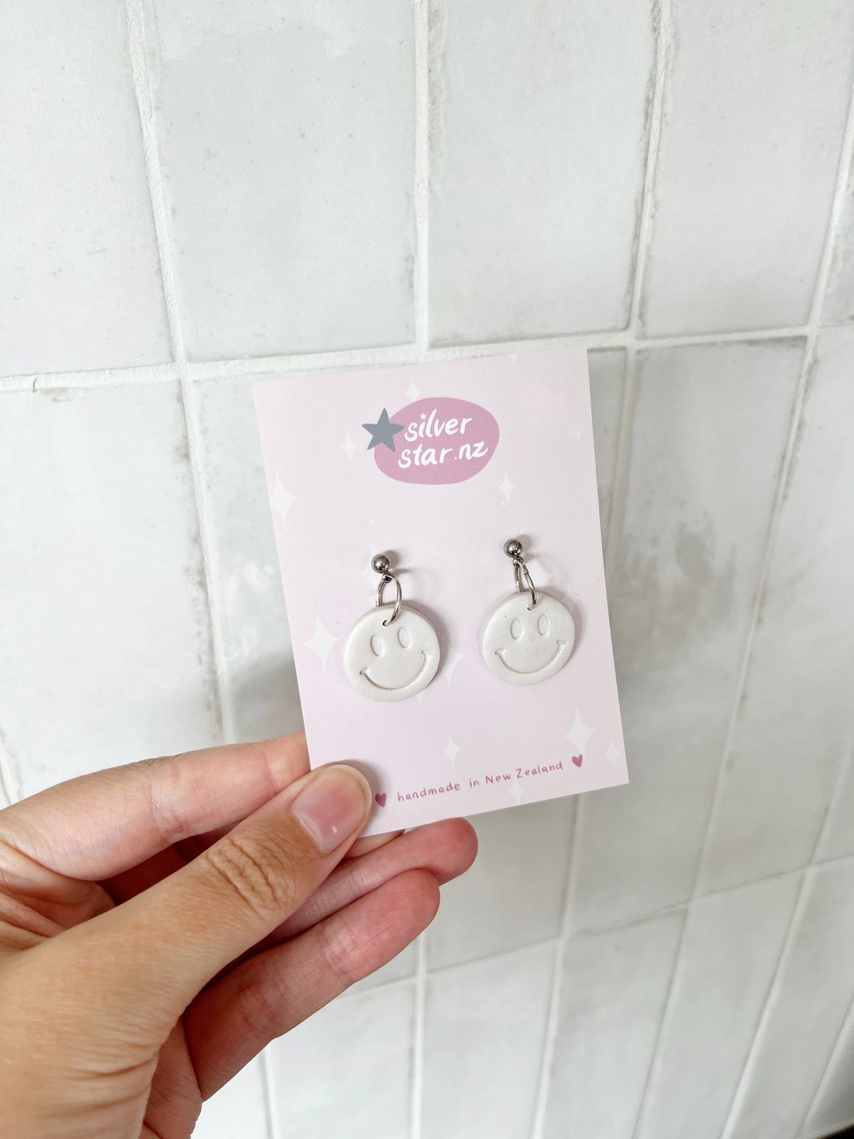 A hand holds a card with “silverstar.nz” showing Smiley Polymer Clay Earrings—white, handmade with dangling smiley faces and stainless steel hooks. The background features white tiles.
