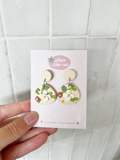 A hand holds a card reading “silverstar.nz” with Floral Organic Oval Polymer Clay Earrings in pastel tones and stainless steel hooks, handmade in New Zealand, shown against a tiled background.