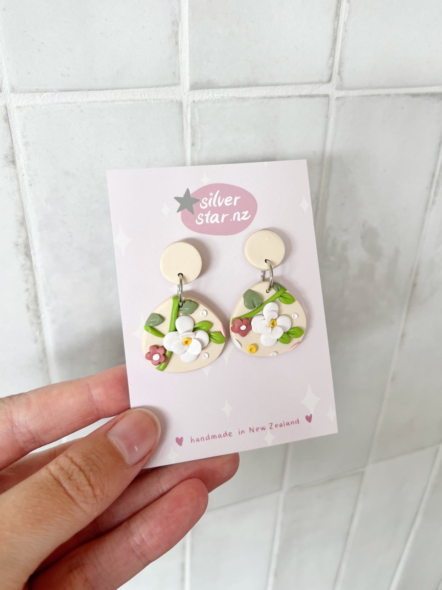 A hand holds a silverstar.nz card displaying Floral Organic Oval Polymer Clay Earrings—cream-colored with white and pink flowers, handmade and attached to stainless steel hooks—against a pale pink background.