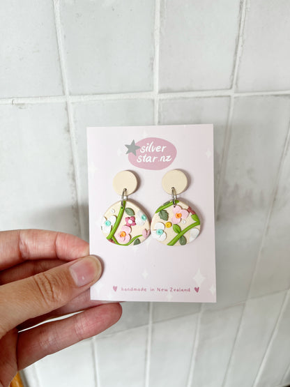 A hand holds a card with Floral Organic Oval Polymer Clay Earrings by silverstar.nz, featuring pastel pink and green details, stainless steel hooks, and handmade in New Zealand text, against a white tiled background.