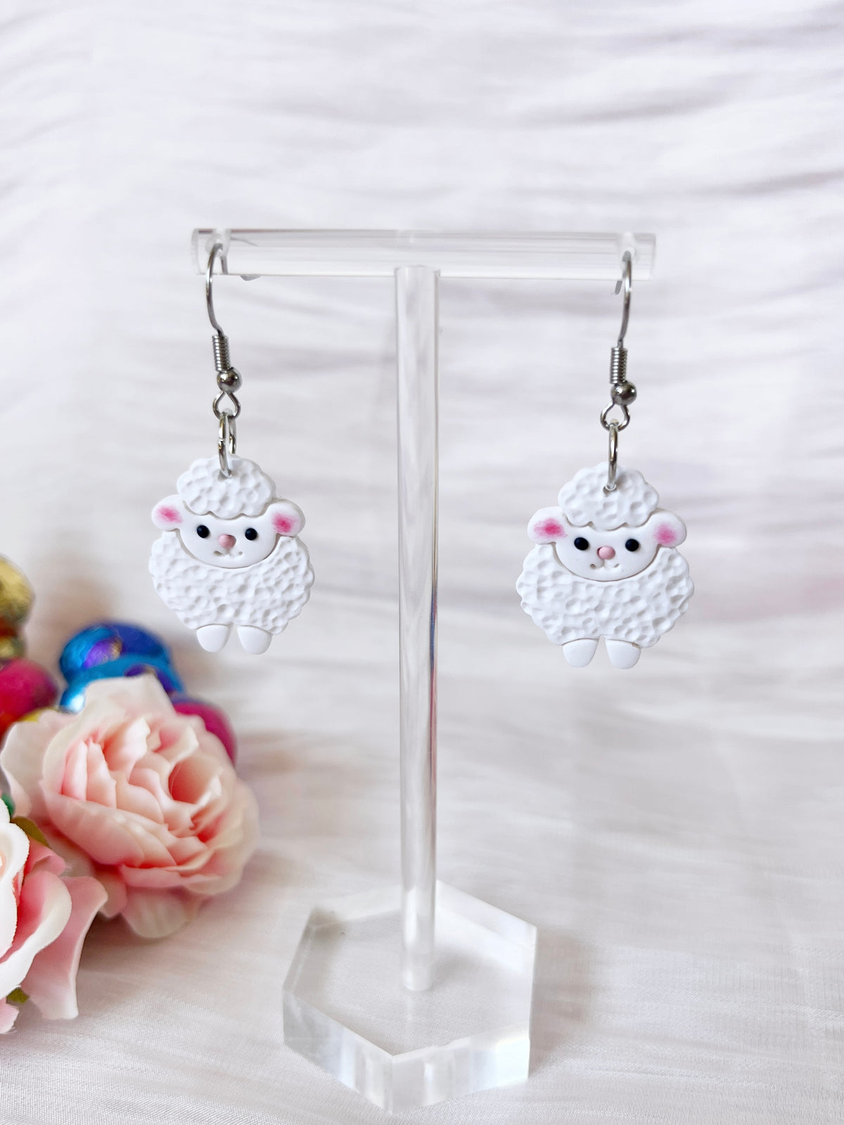 Fluffy Sheep Polymer Clay Earrings by silverstar.nz dangle on a transparent stand, featuring sheep with pink cheeks and black eyes. Pink roses and colorful wool balls enhance the unique design, all set against a soft white fabric backdrop.