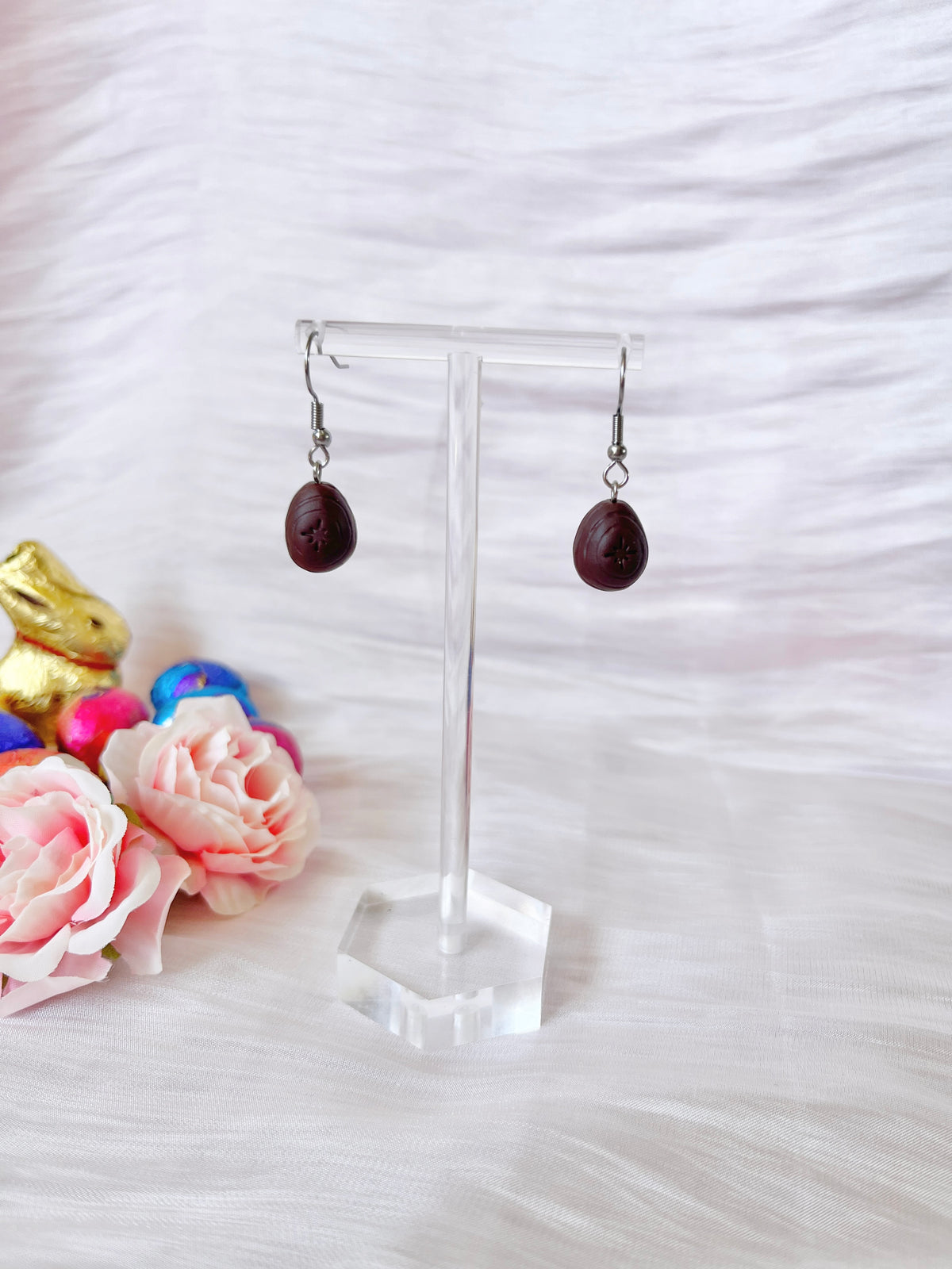 The Mini Creme Egg Polymer Clay Earrings by silverstar.nz are showcased on a clear stand, surrounded by pink roses and colorful chocolate bunnies against textured white fabric. These handmade earrings feature a unique, charming, and whimsical design.