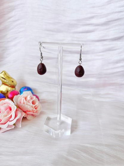 The Mini Creme Egg Polymer Clay Earrings by silverstar.nz are showcased on a clear stand, surrounded by pink roses and colorful chocolate bunnies against textured white fabric. These handmade earrings feature a unique, charming, and whimsical design.