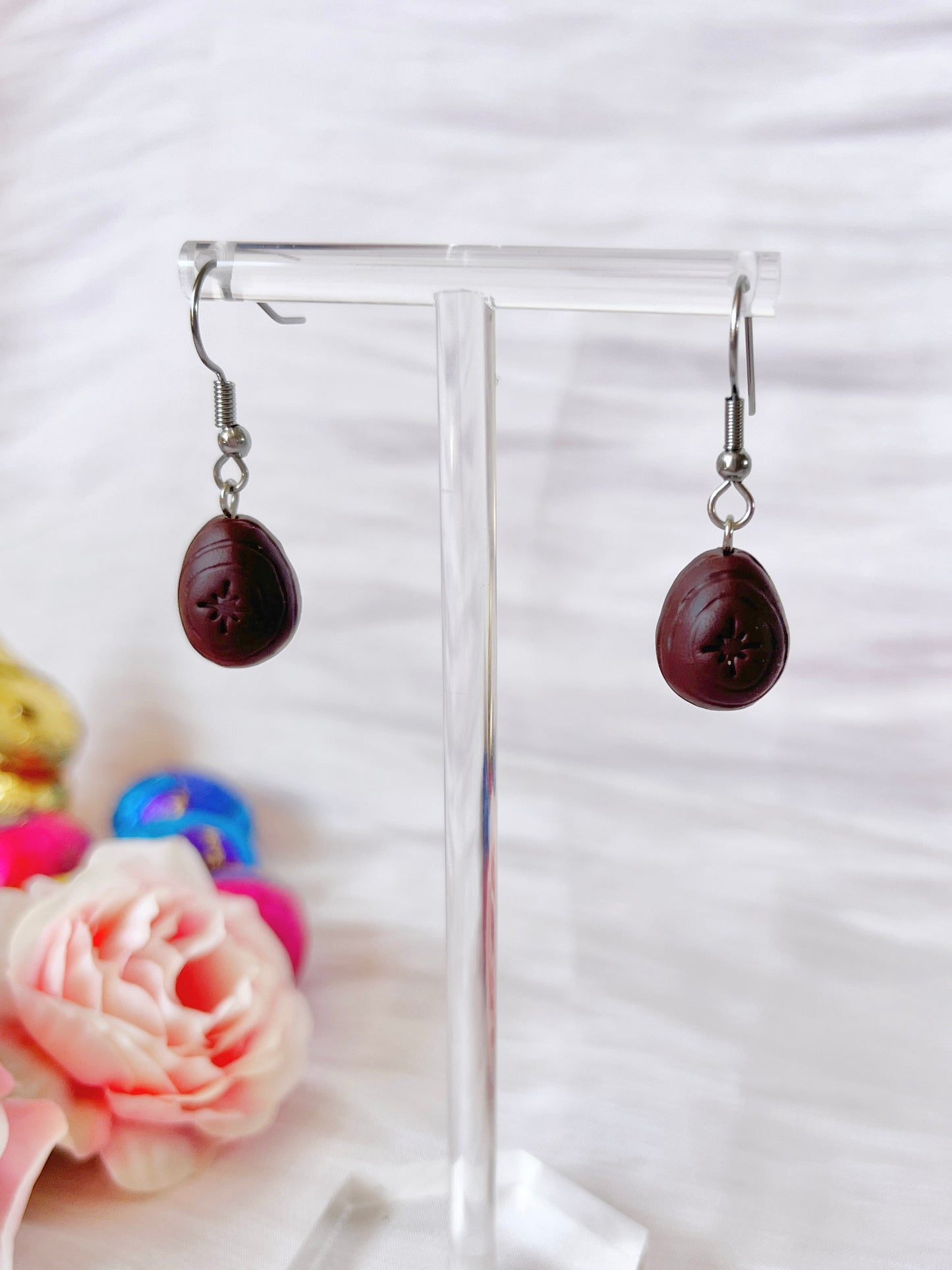 Close-up of silverstar.nzs unique Mini Creme Egg Polymer Clay Earrings hanging on a transparent display stand. A soft pink rose and colorful blurred objects in the background provide a gentle, pastel contrast to these handmade earrings.