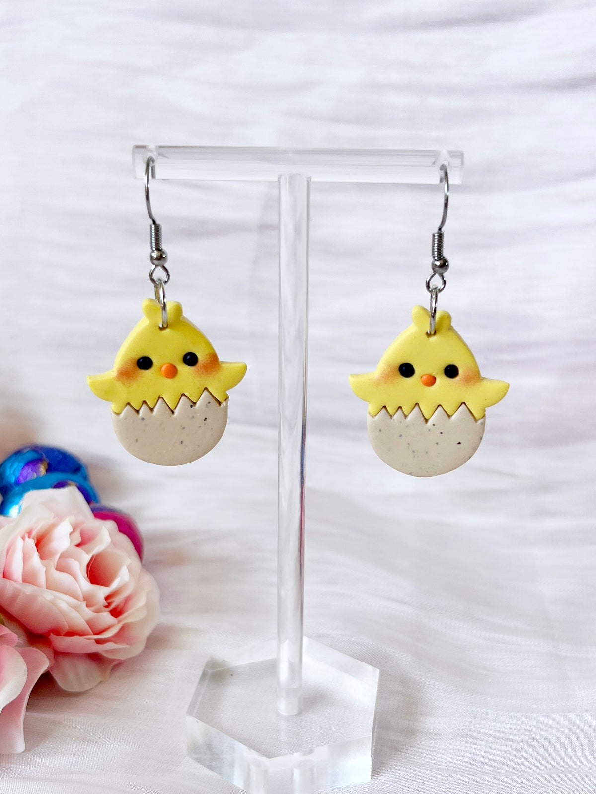 The Egg Baby Chick Polymer Clay Earrings by silverstar.nz feature a unique design of yellow chicks with small wings, orange beaks, and black eyes hatching from eggs on a clear stand. Pink roses and colorful fabric are in the background.