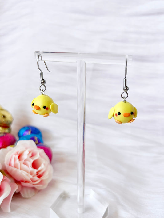 A pair of silverstar.nzs Mini Baby Chick Polymer Clay Earrings, featuring yellow chicks with orange beaks and pink cheeks, hang on a clear stand. A pink rose and colorful yarn balls enhance their charm against a white textured background.