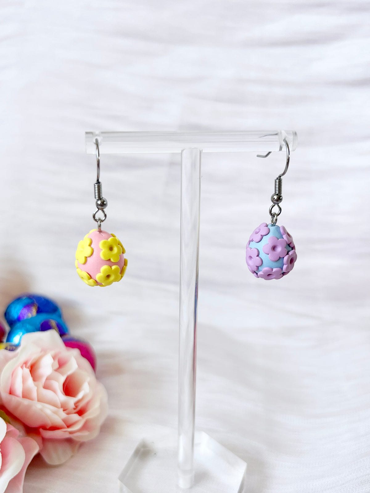 The Floral Egg Polymer Clay Earrings by silverstar.nz are displayed on a clear stand, featuring one pink earring with yellow flowers and another purple earring with lilac flowers, both crafted from polymer clay. Stainless steel hooks complete the set, against a soft focus backdrop of pink roses and colorful eggs.