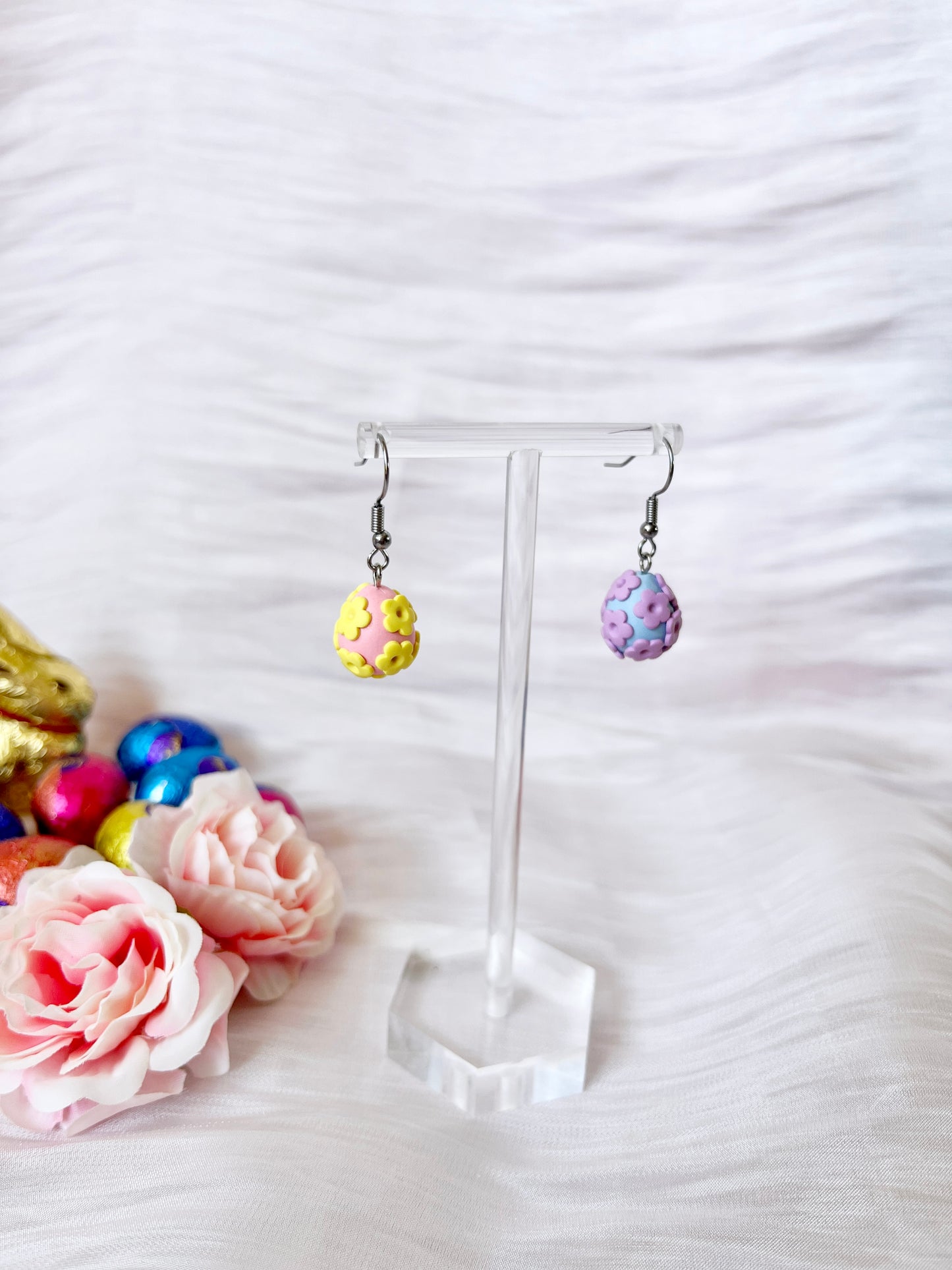 The Floral Egg Polymer Clay Earrings from silverstar.nz feature pastel egg shapes, one yellow with flowers and the other purple, crafted by hand with polymer clay. They come with sturdy stainless steel hooks and are displayed on a clear stand against a soft backdrop surrounded by faux flowers and colored eggs.