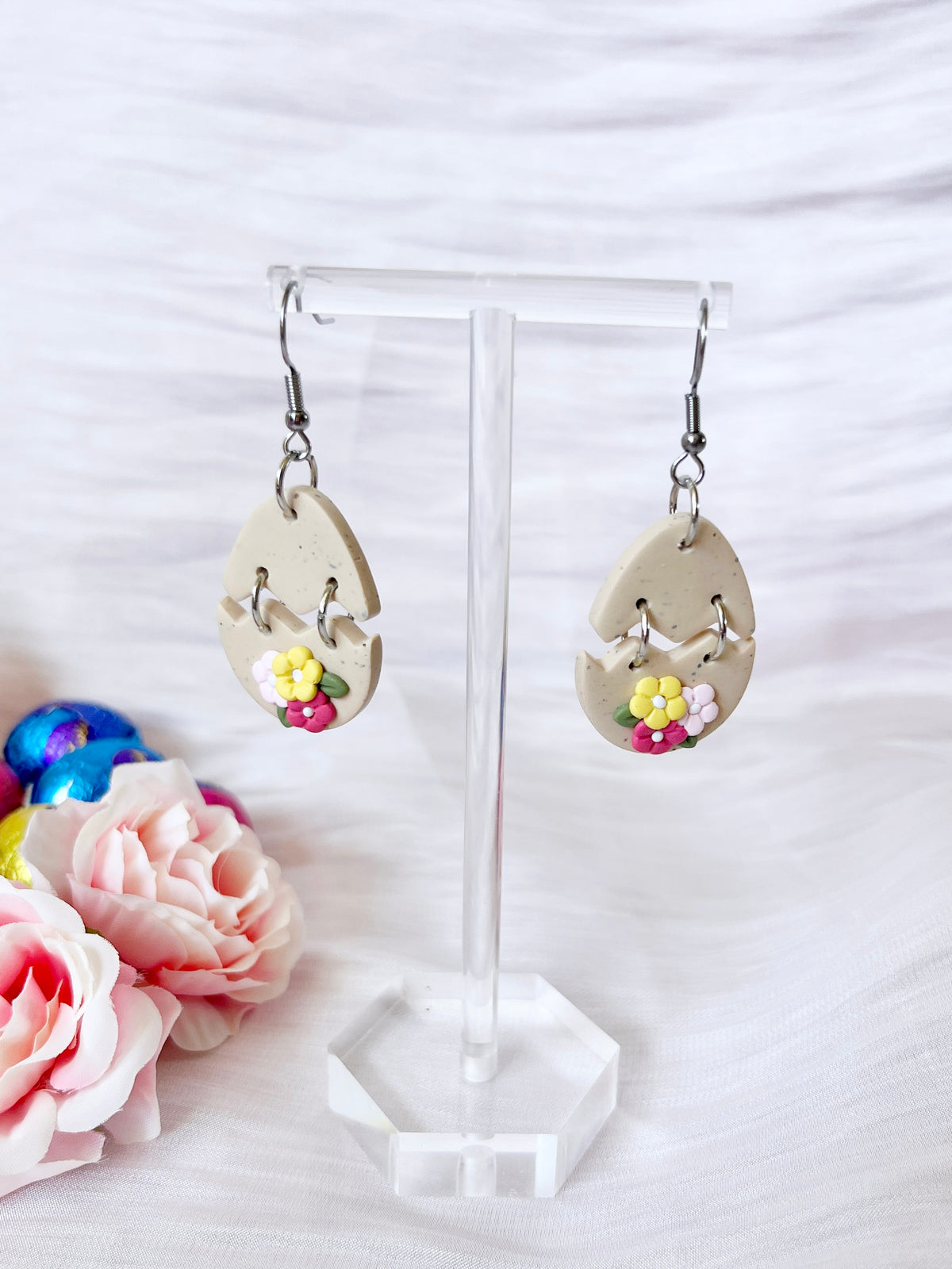 Broken Egg Polymer Clay Earrings by silverstar.nz feature a beige base with vibrant floral designs, elegantly displayed on a clear acrylic stand. Soft pink roses and colorful beads decorate the white textured backdrop, complemented by stainless steel hooks for lasting wear.