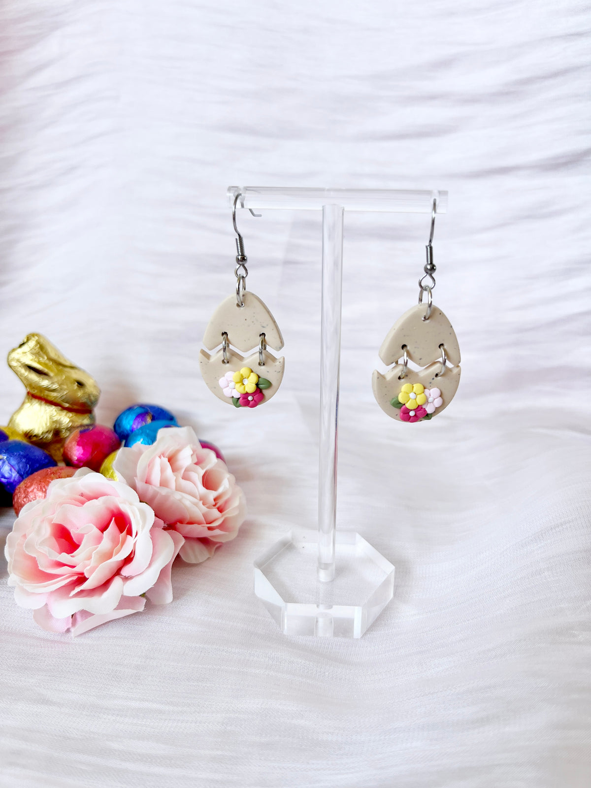 The Broken Egg Polymer Clay Earrings by silverstar.nz, teardrop-shaped and handmade with floral details, are elegantly presented on a stand. In the background, colorful chocolate eggs and a gold bunny figure are complemented by pink artificial roses against a white textured backdrop.