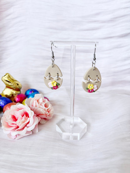 The Broken Egg Polymer Clay Earrings by silverstar.nz, teardrop-shaped and handmade with floral details, are elegantly presented on a stand. In the background, colorful chocolate eggs and a gold bunny figure are complemented by pink artificial roses against a white textured backdrop.