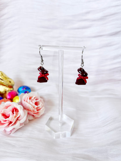 Chocolate Bunny Polymer Clay Earrings by silverstar.nz hang on a transparent stand against a white, wavy fabric background. Pink roses and colorful Easter eggs adorn the left side, enhancing the funky charm of these handmade treasures.